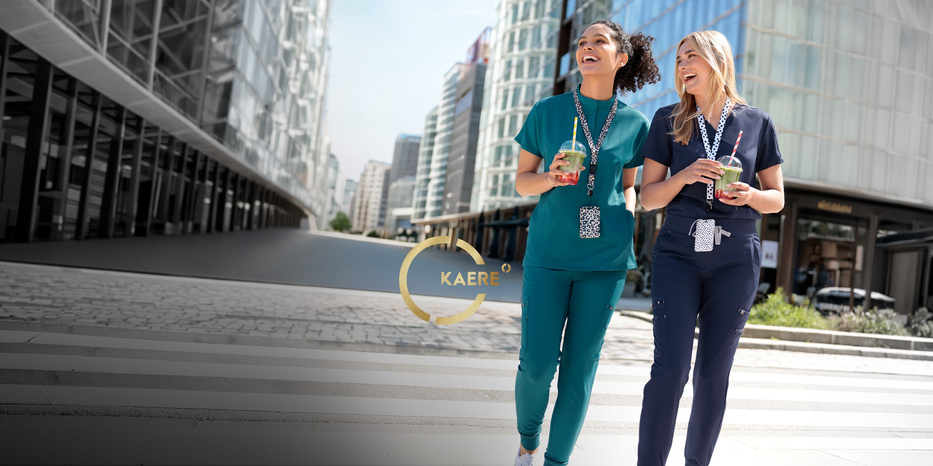 Twee vrouwen in turquoise en donkerblauwe KAERE werkkleding met drankjes in een moderne stedelijke omgeving.